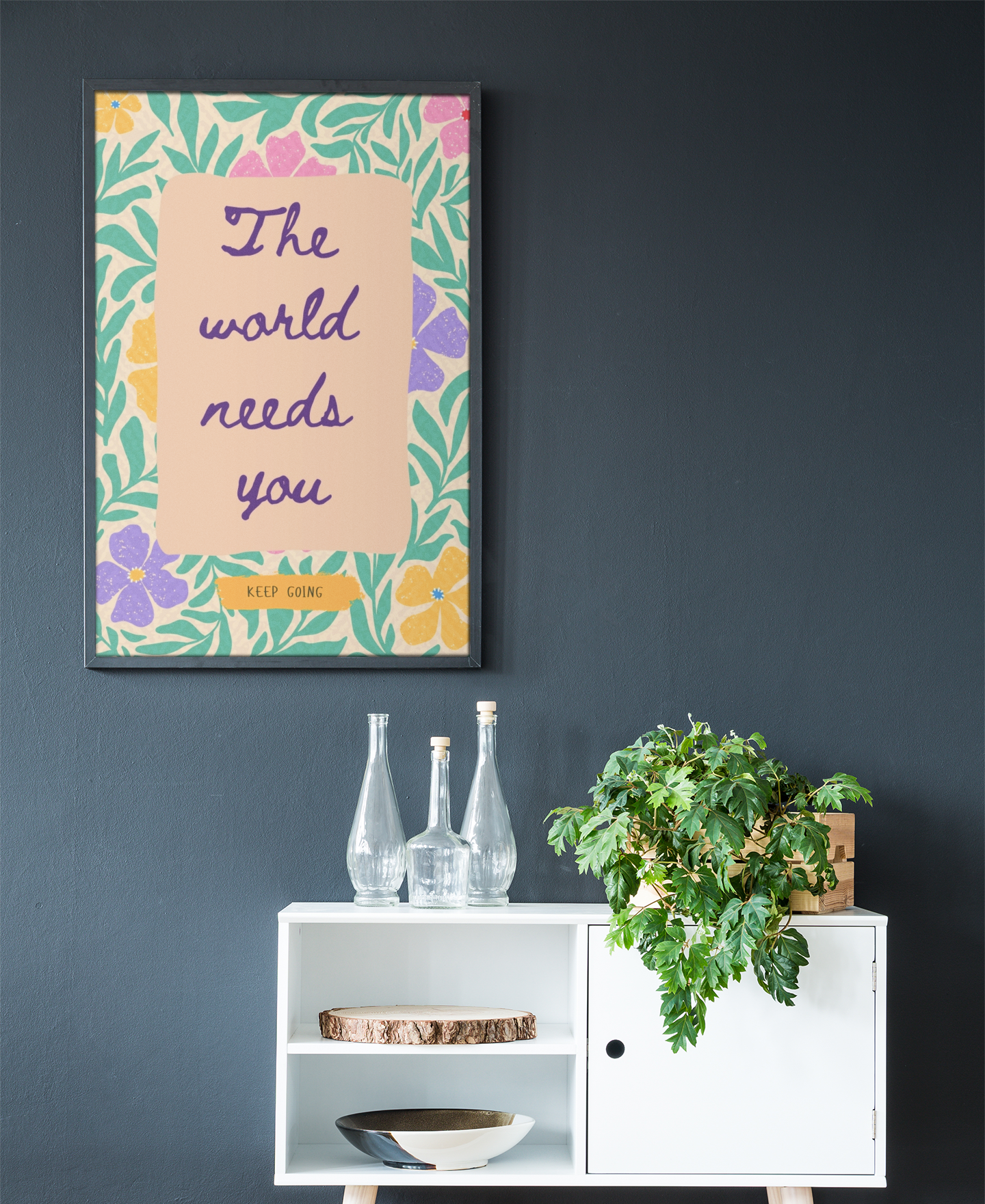 A framed art print with a colorful background and text that reads 'The world needs you', displayed on a dark wall above a white side table with a plant and glassware.