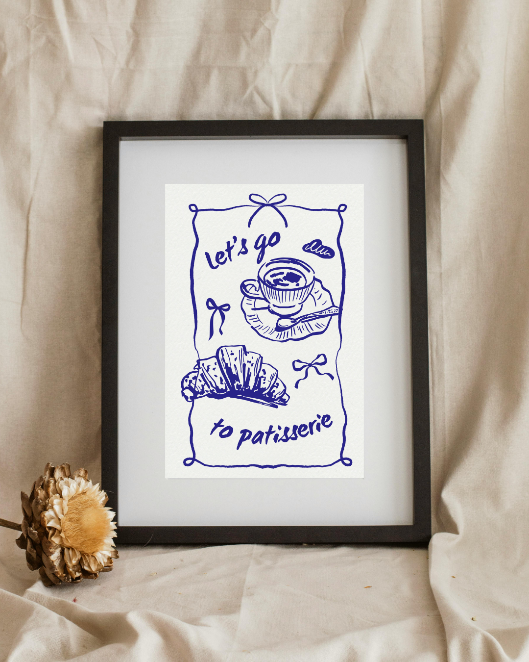 Framed artwork with blue line drawing and text 'Let's go to patisserie' on a beige fabric background.